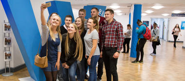 A large group of students stand together smiling in main reception, while the student at the front takes a selfie with her mobile phone.