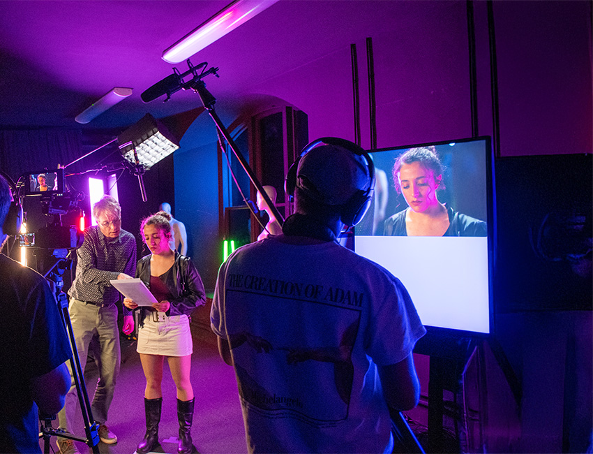 Students with camera and sound equipment. A student stands in front of the camera looking at a script with lecturer. 