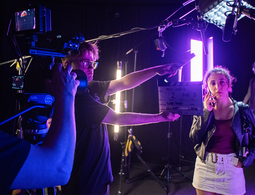 A camera points at a clapperboard, behind it stands a student on the phone, lit up in neon light