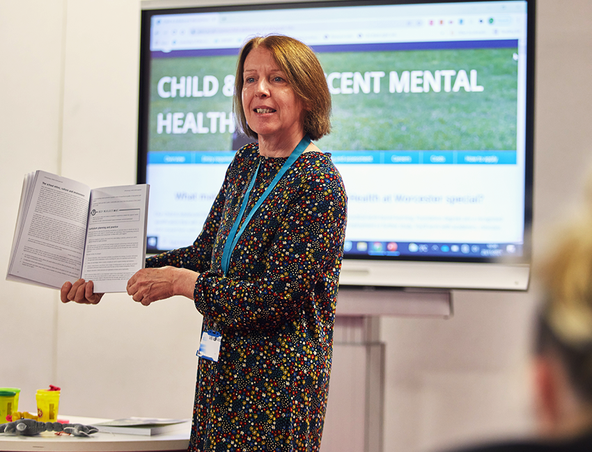 Person stood in front of a large screen, showing an open book as she talks to an audience