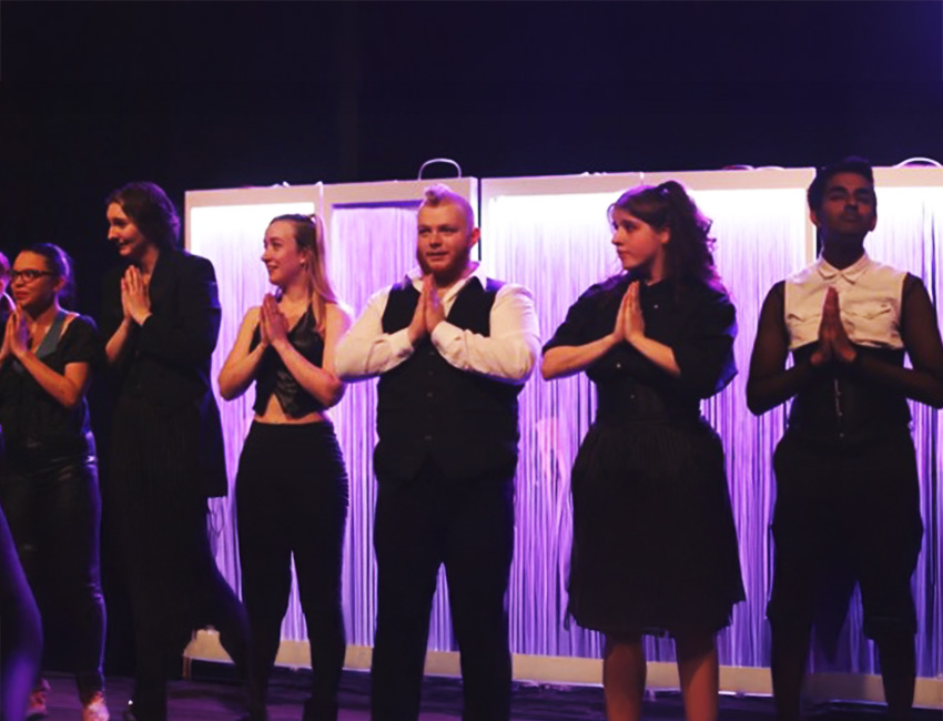 Students lined up on stage wearing black and white clothes, lit up with neon purple light with a white curtain behind