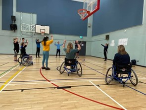 Mixed dance class in gym hall with three students in wheelchairs led by lecturer standing in front of the group