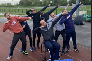 Six men standing behind a man in a wheelchair on a running track all 'dabbing'