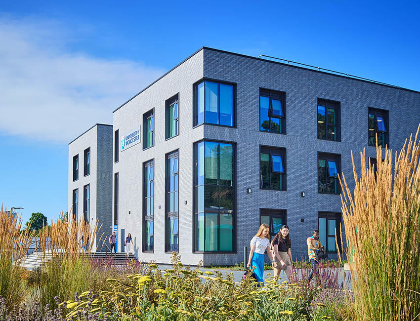 A modern grey-brick university building with large glass windows on a bright sunny day. Tall grasses and colourful plants fill the landscaped foreground, while a few people walk along the path beside the building.