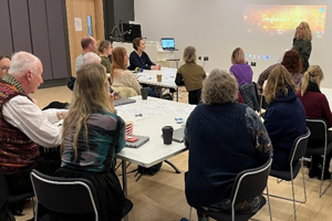 Organiser of the Transformative Tales event at the front of the room delivering a session to attendees grouped at tables.