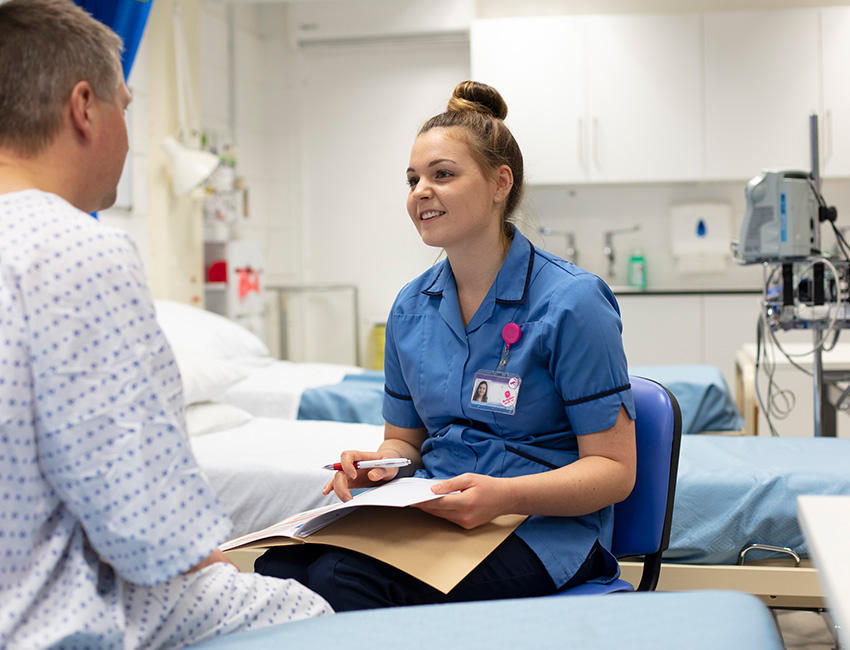 A nurse in a blue uniform sits in a simulation room holding a clipboard and pen while speaking with a patient who is seated on a bed. Medical equipment and additional beds are visible in the background.
