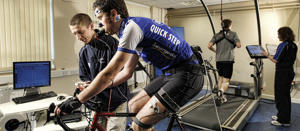 A sports analyst takes scientific readings from a man on a stationary bicycle. The man on the bicycle has measuring equipment strapped to his face.