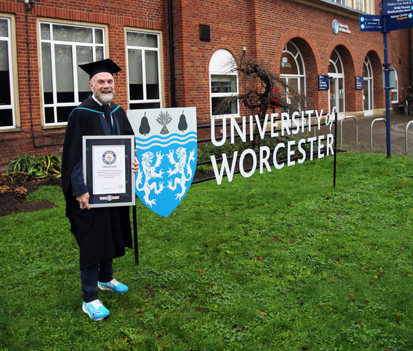 Chris poses in cap and gown by the University of Worcester sign on campus