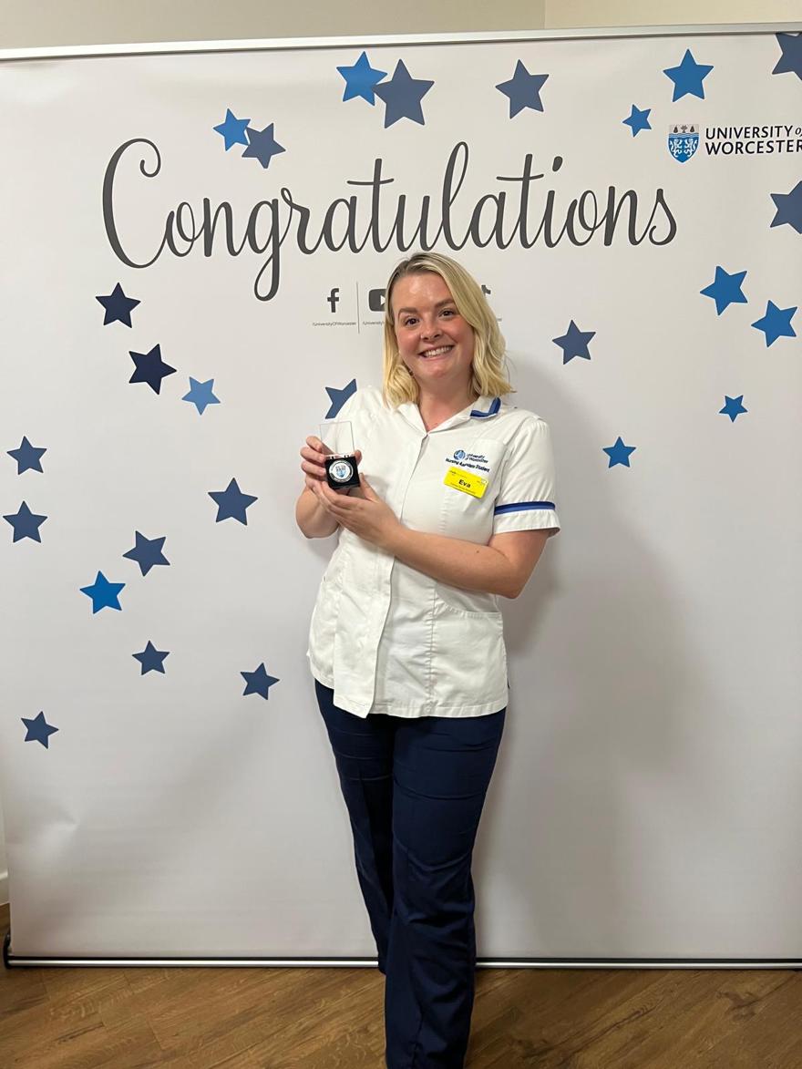 Eva Savage poses in front of a banner which says 'congratulations' while holding an award