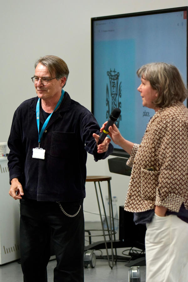 John Cussans and Desdomona McCannon address a crowd with a microphone in front of a screen