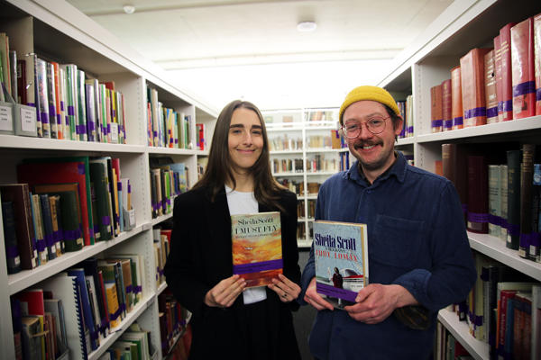 Jasmine Kee and Tom Mandall pose with books between bookcases in a well lit library