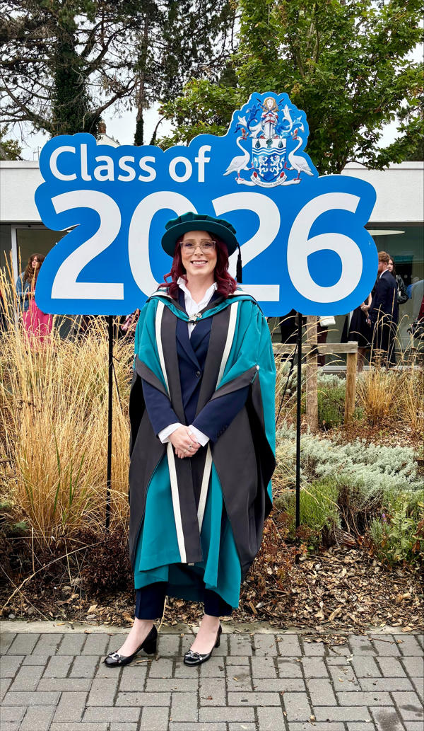 A full body portrait of a graduate in cap and gown in front of a sign reading 'class of 2026'