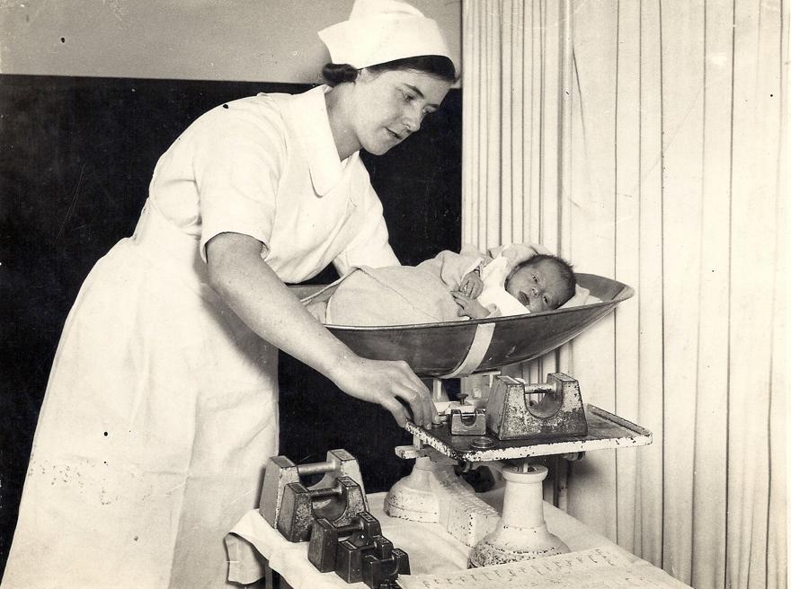 A black and white image of a midwife weighing a baby in traditional scales