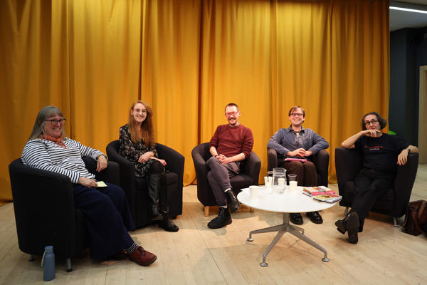 Five people sit in chairs in a semi circle in front of a yellow curtain