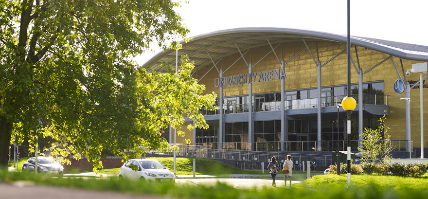 A gold arena building with a domed roof, surrounded by leafy trees on a bright and sunny day.