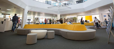 Groups of students walking around and sitting on modern yellow and grey sofas in a large, open study space.