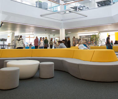 Groups of students walking around and sitting on modern yellow and grey sofas in a large, open study space.
