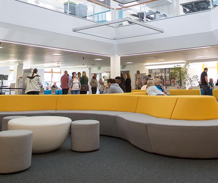 Groups of students walking around and sitting on modern yellow and grey sofas in a large, open study space.