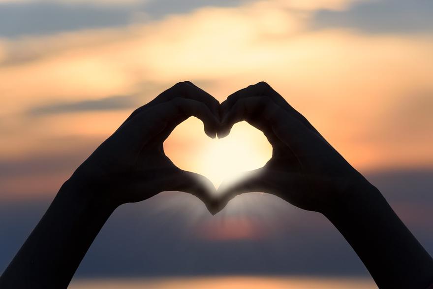 A black silhouette of two hands forming a heart shape in front of a beautiful sunset backdrop.