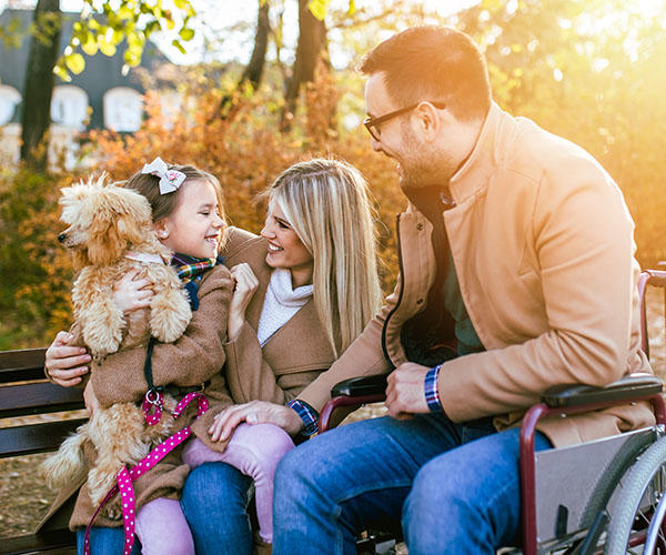Two adults, one of whom is in a wheel chair, are talking to a child. The child is smiling and holding a teddy bear