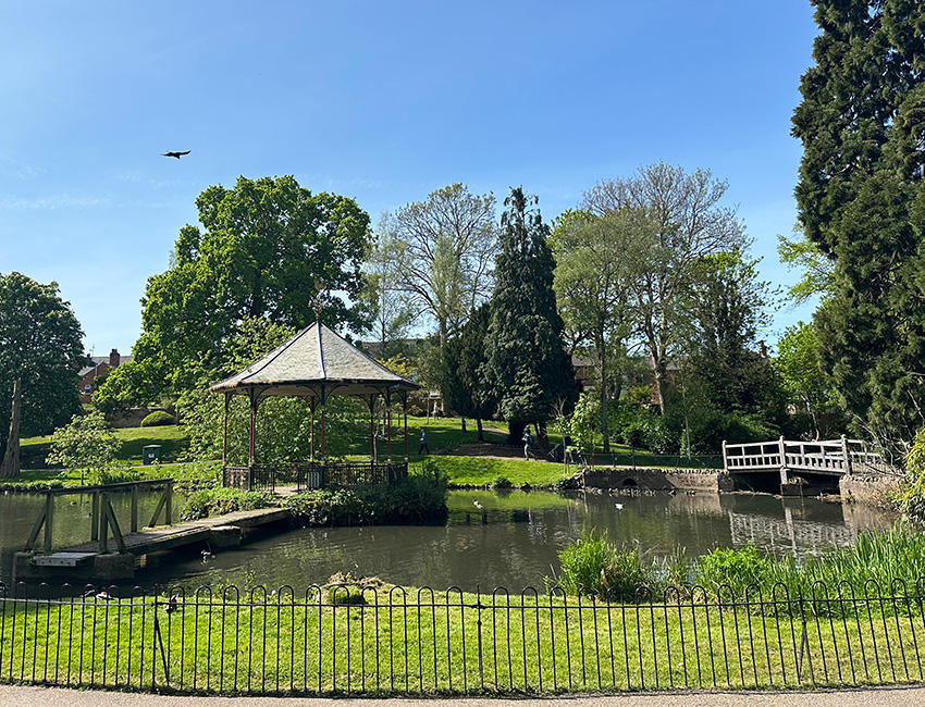 A band stand in the middle of a large pond with a curved wooden bridge to the right