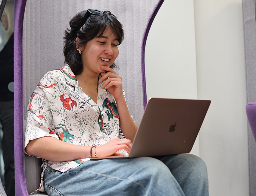 A girl sat in a study area smiling at her laptop