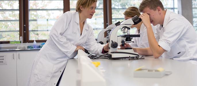 Two students looking into their microscopes whilst the lecturer leans over the lab counter to talk to them.