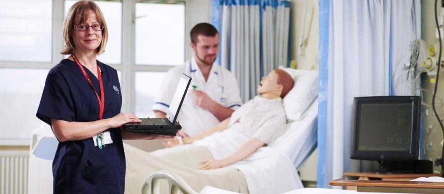 Nurses stood next to mannequin in hospital bed.