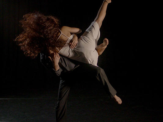 Male dancer dressed in black holding a female dancer in white dress, with a black background