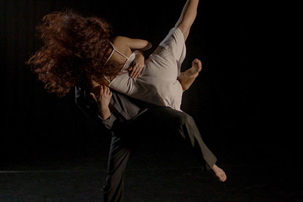 Male dancer dressed in black holding a female dancer in white dress, with a black background