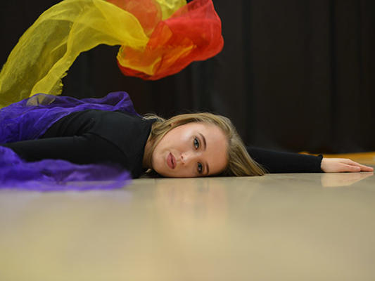 Female student lying on the floor, facing camera with multi-coloured scarves