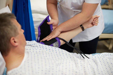 Man in hospital gown laying down on hospital bed, with a blood pressure monitor being strapped onto his arm by a nurse