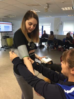 A female Occupational Therapy trainee is applying a blood pressure monitor to a female student's arm as a practical on our occupational therapy degree.