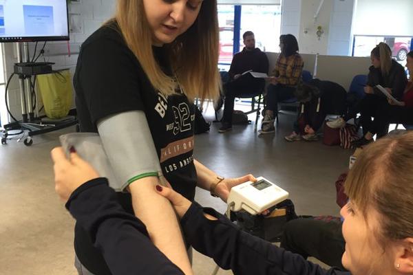 A female Occupational Therapy trainee is applying a blood pressure monitor to a female student's arm as a practical on our occupational therapy degree.
