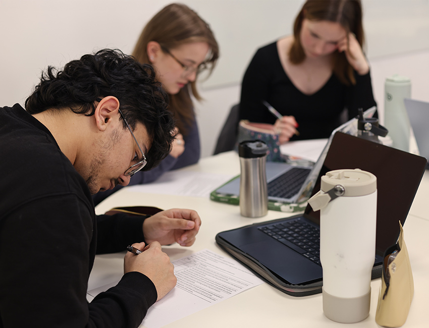 A male student writing on a notebook with two female students in the background