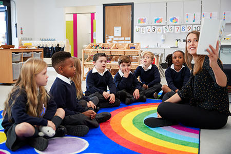 A group of students are being read a story by a teacher sitting on the floor of a classroom next to them.