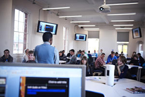 a lecture room with screens on the wall a lecturer at the front and students at their desks