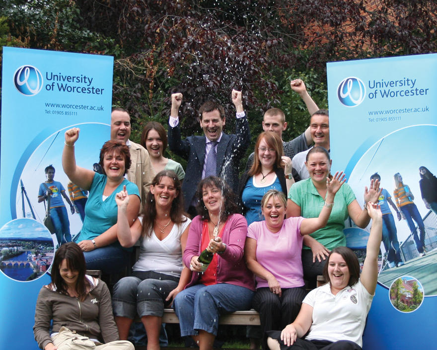 University of Worcester students and staff celebrating with champagne and raised handsbetween two university banners.