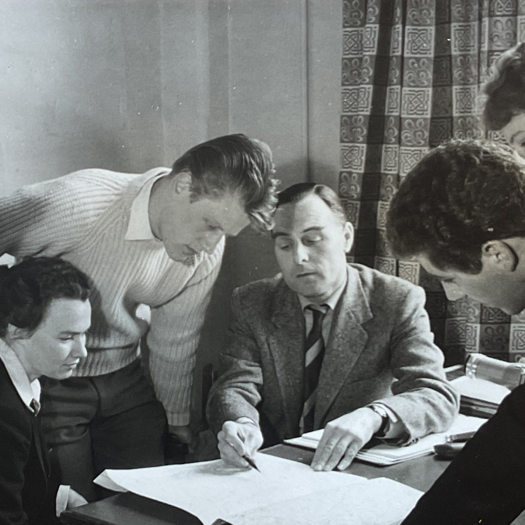 A group of five men gathered around a table reviewing large documents