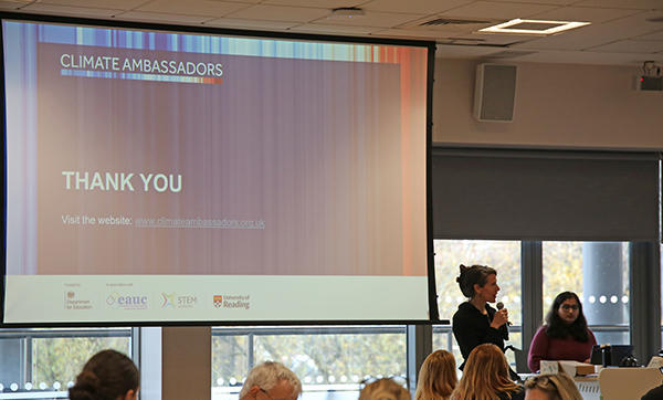 A projector screen displays a thank you message from the Climate Ambassadors and one person is standing next to the screen holding a microphone while others are watching.