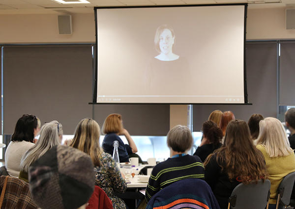 Jess Phillips MP, Minister for Safeguarding, is visible on a projector screen delivering a message to a group of people, who are all looking at the screen.