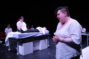 Students acting out a scene in a hospital. One student close to the camera looks on in anguish, while another lies on a bed behind with a student placing a sheet over them.