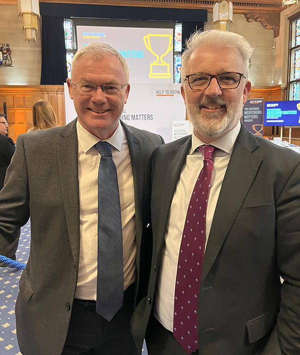 Two men in suits stood side by side looking at the camera, smiling