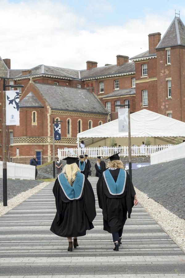 Two people wearing graduation robes photographesd from the back walking up a path with a large red brick building in the background