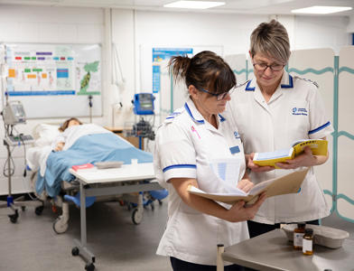 Two nurses in white uniforms review paperwork and a reference book in a clinical skills training room. A patient lies in a hospital bed in the background, surrounded by medical equipment and screens.
