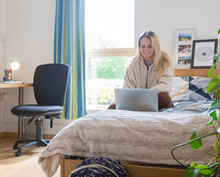 a smiling student is sitting on a bed in University accommodation