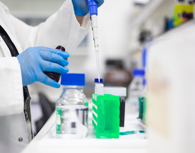 A close up of a student using a long pipette wearing gloves and lab coat, with chemicals on table