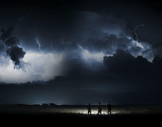 Three people stand silhouetted in a field at night as lightening strikes the sky