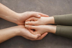 Two pairs of hands gently holding each other on a gray surface, symbolizing support and care.