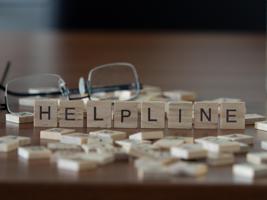 Wooden letter tiles arranged to spell 'HELPLINE' on a table with scattered tiles around and a pair of eyeglasses in the background.